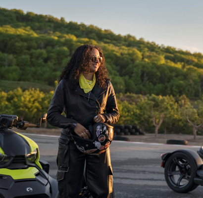 women with long curly hair dressed in street riding gear, holding a helmet on a road with her spyder. 